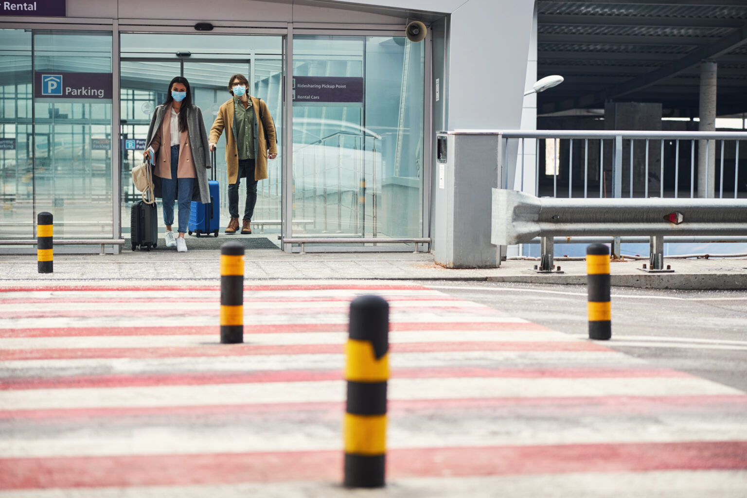 Stylish lady and gentleman with travel suitcases wearing protective face masks while heading to pedestrian crossing at airport